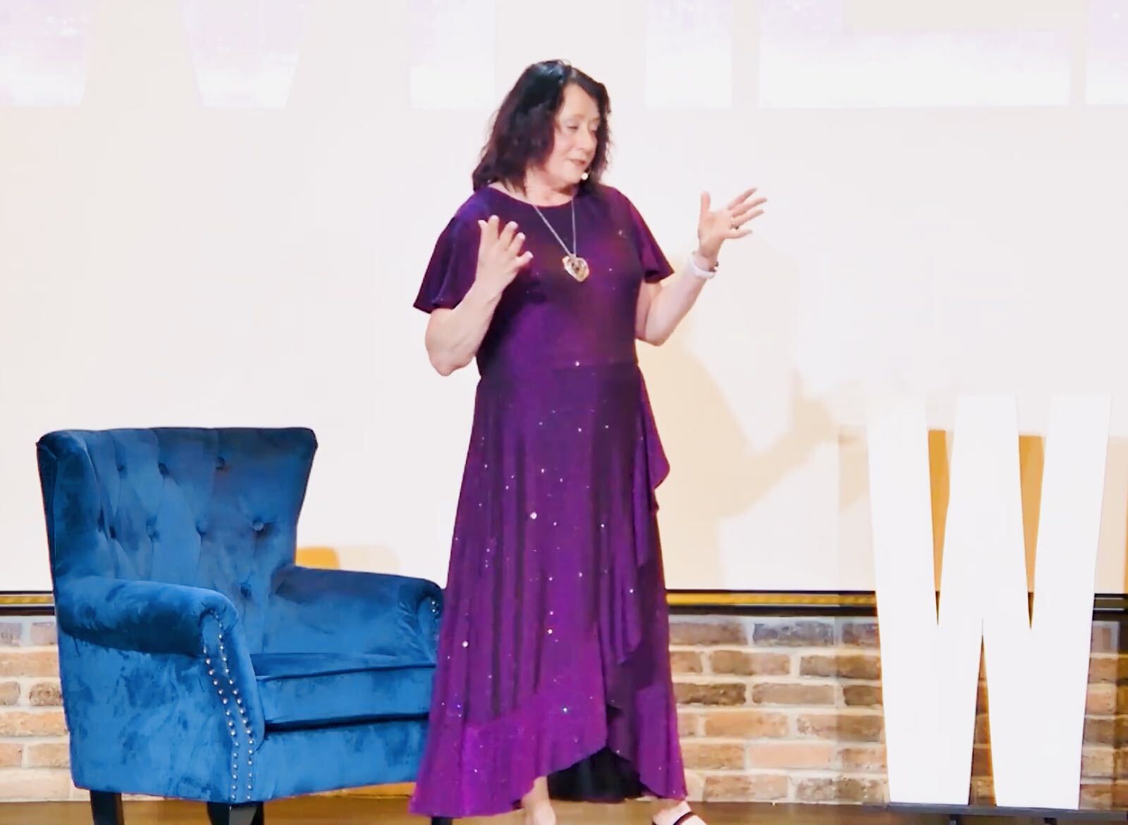 Rhonda Deaver speaking on stage in a purple gown with her hands extended